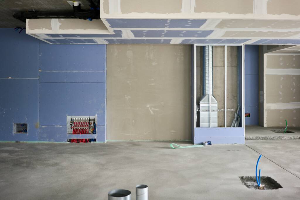 Interior view of a building under construction, featuring exposed wiring, unfinished drywall, ductwork, and conduits emerging from a concrete floor.
