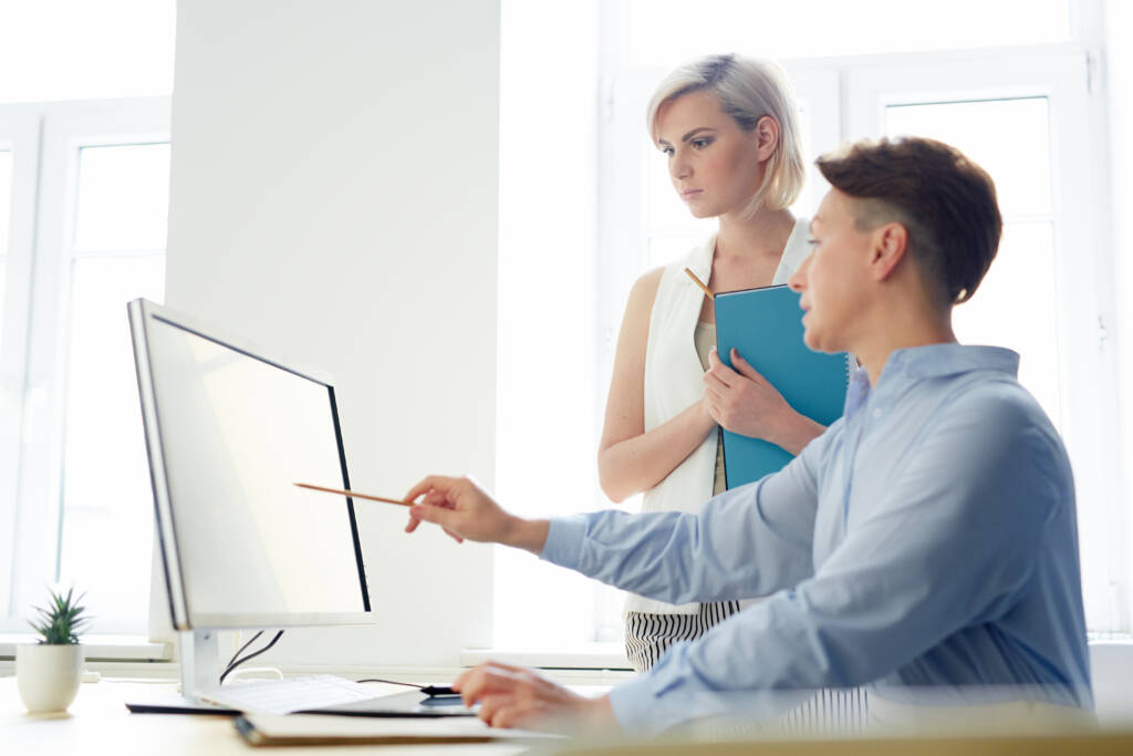 A man and woman review content on a desktop monitor in a bright office setting.